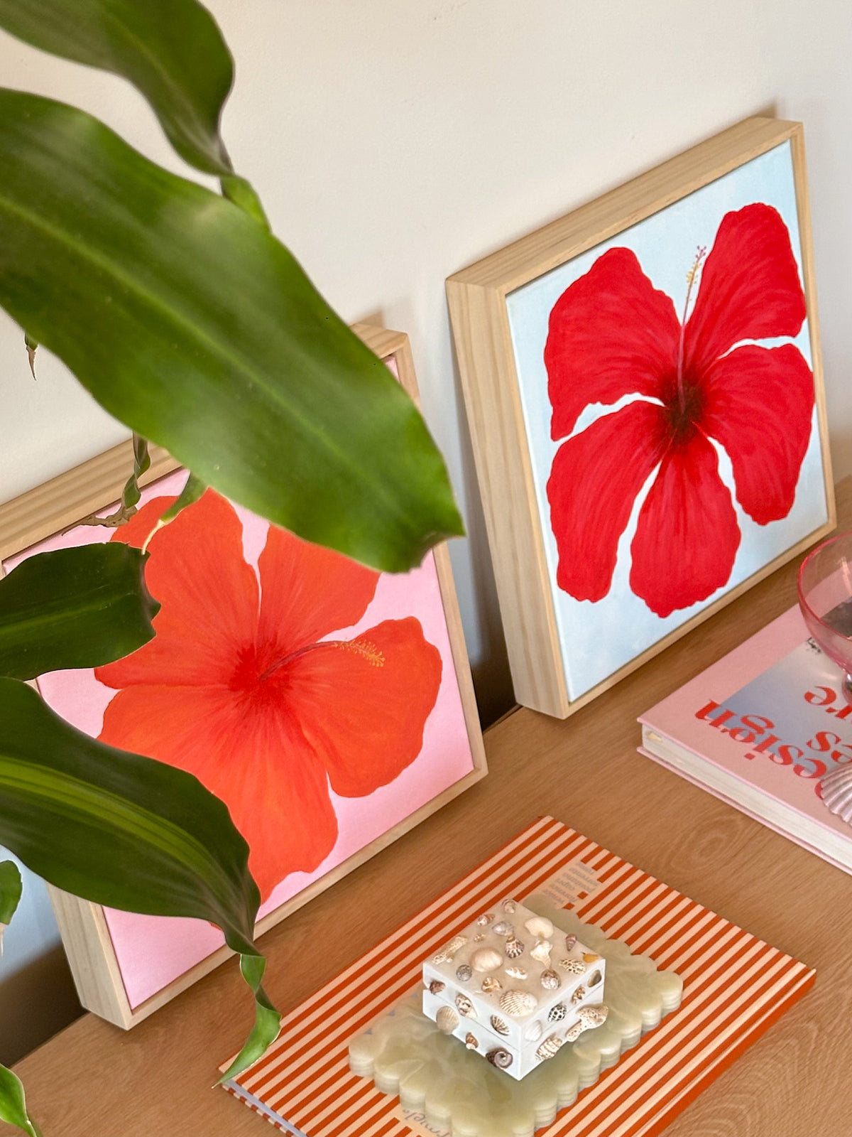 Two framed original artworks of red & orange hibiscus flowers on a wooden surface with a plant and decorative items.
